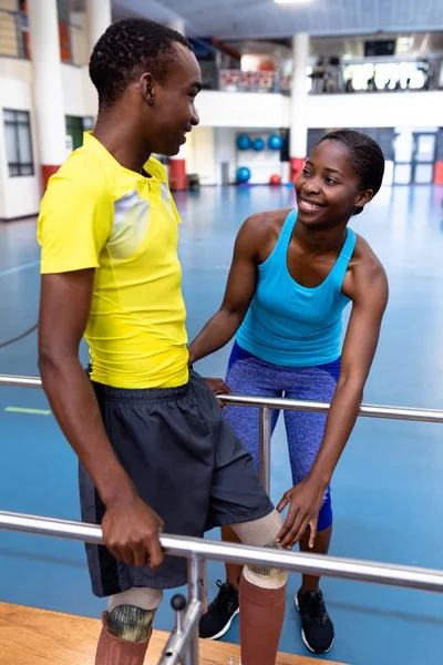 幸せアフリカ系アメリカ人女性理学療法士のフロントビュー障害者のアフリカ系アメリカ人男性がスポーツセンターで平行棒で歩くのを助けます — ストック写真