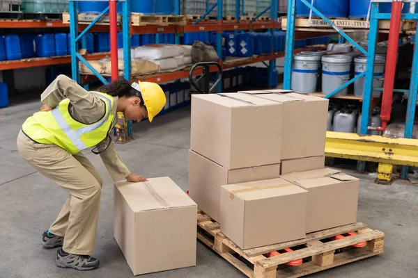 Female staff suffering from back pain while holding heavy cardboard box in warehouse.