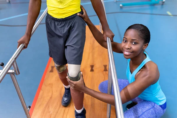 アフリカ系アメリカ人の理学療法士の肖像障害者がスポーツセンターの平行棒で歩くのを助ける — ストック写真