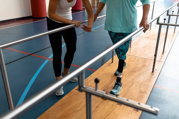 スポーツセンターでパラレルバーで歩く障害のある女性を助ける女性理学療法士の低いセクション — ストック写真