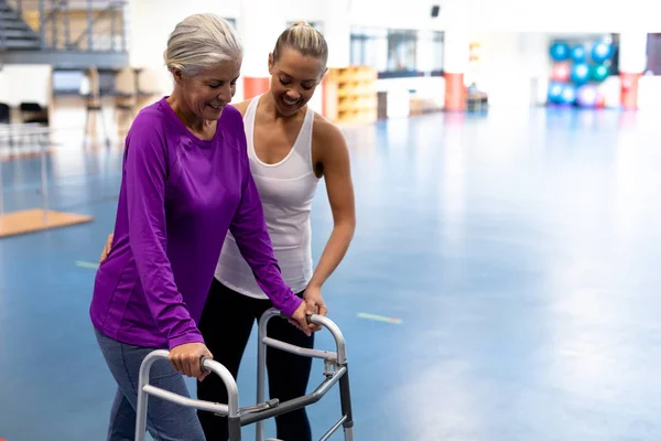 障害のあるシニア女性がスポーツセンターでウォーカーと歩くのを助ける女性理学療法士の側面図 — ストック写真