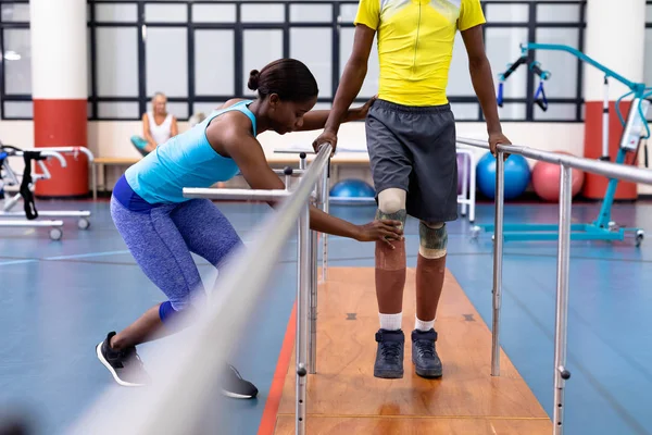 アフリカ系アメリカ人女性理学療法士のサイドビュー障害者がスポーツセンターで平行棒で歩くのを支援 — ストック写真