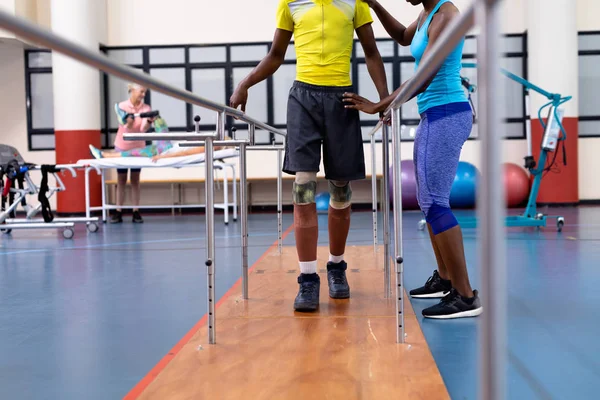 障害者がスポーツセンターで平行棒で歩くのを助ける理学療法士の低いセクション — ストック写真