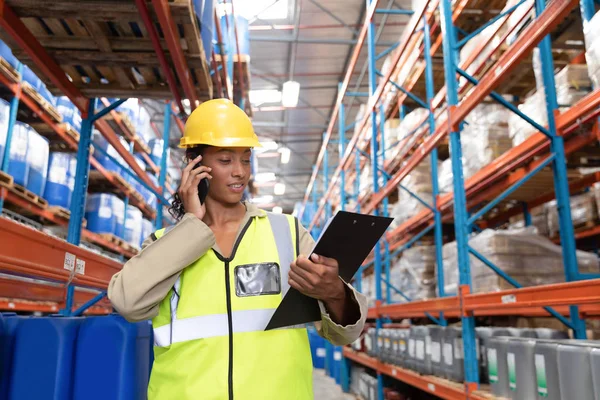 倉庫で携帯電話で話しながらクリップボードを見ている女性労働者の正面図 — ストック写真