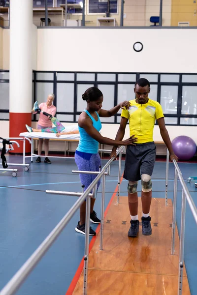 アフリカ系アメリカ人女性理学療法士のフロントビューは 障害のあるアフリカ系アメリカ人男性がスポーツセンターで平行棒で歩くのを助ける — ストック写真