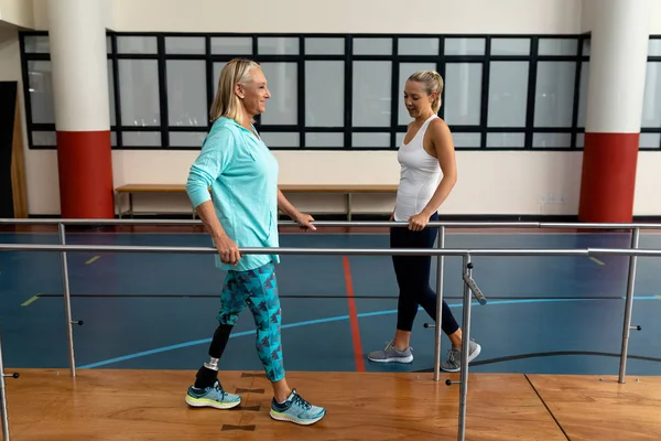 スポーツセンターでパラレルバーで歩く障害のあるシニア女性を助ける女性理学療法士の側面図 — ストック写真