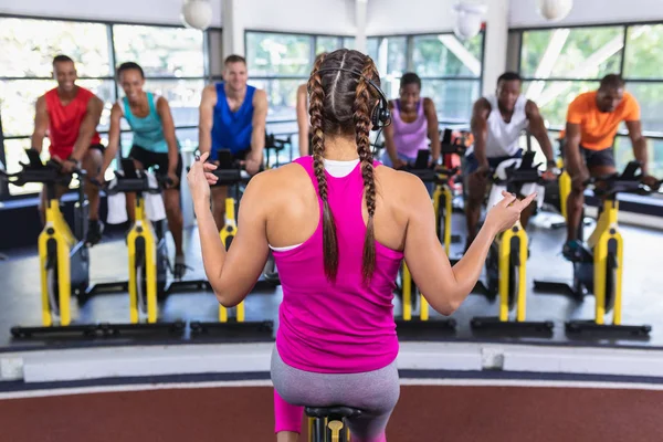 白人女性トレーナーの背面ビューフィットネスセンターで運動自転車に取り組むために人々を訓練します 健康的な人々が働いているとスピンクラスでトレーニングと明るい現代のジム — ストック写真