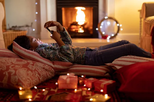 圣诞节时 一个年轻的混血女孩坐在客厅里 躺在地板上的垫子上 用智能手机 — 图库照片