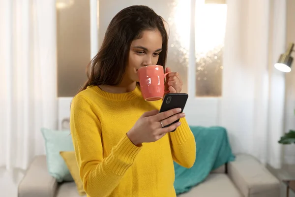 前视特写一个快乐的年轻白种妇女喝一杯咖啡 微笑着看着智能手机在她的客厅 — 图库照片