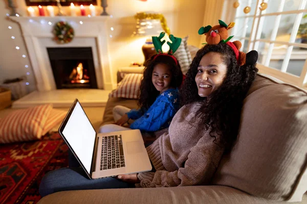 クリスマスに幼い娘とソファに座り ノートパソコンを使い お祝いの帽子をかぶったカメラに微笑む混合人種女性の肖像画 — ストック写真