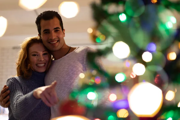一个年轻的混血男子和一个年轻的白种妇女在家里装饰的圣诞树旁微笑和拥抱的正面视图 — 图库照片