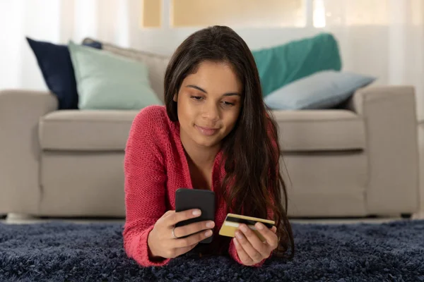 前视特写一个快乐的年轻白人妇女使用智能手机 并拿着她的信用卡躺在家里的客厅地板上 — 图库照片