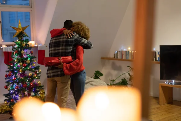 飾られたクリスマスツリーの横のアパートの居間で抱き合う若い男女の側面図 プレゼントを持っている女性 — ストック写真