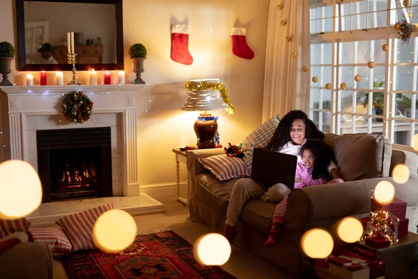 圣诞节时 一位混血女人和她的小女儿坐在客厅里 坐在客厅里 用笔记本电脑微笑着 前景中闪烁着灯光 — 图库照片