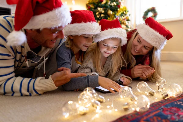 圣诞节时 一对白种人夫妇躺在地板上 带着年幼的儿子和女儿躺在客厅里 戴着圣诞老人的帽子 戴着平板电脑 — 图库照片