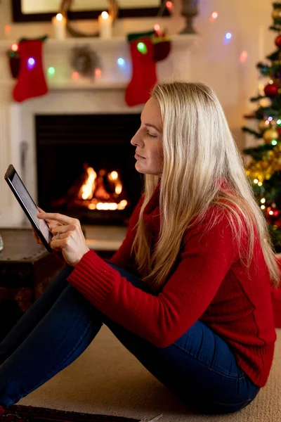 圣诞节时 一位年轻的白种妇女使用平板电脑坐在客厅的壁炉前 — 图库照片