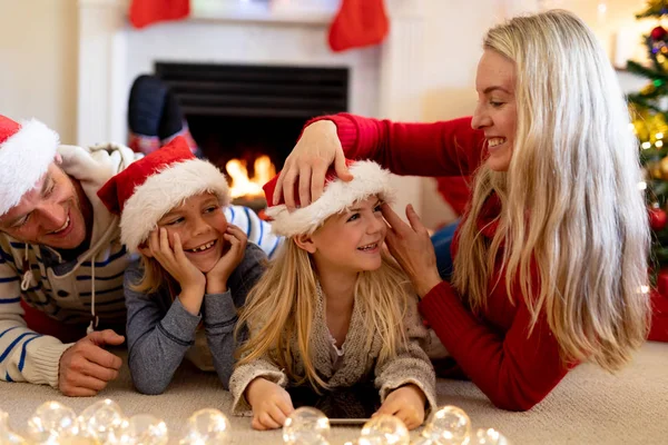 クリスマスの時期に彼らの居間でサンタの帽子の床に横たわっている彼らの若い息子と娘を持つ白人のカップルの正面図 母親は彼女の娘の帽子を調整します — ストック写真