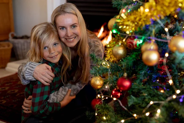 圣诞节时 一位年轻的白种妇女拥抱着坐在客厅地板上的小儿子 旁边是一棵装饰的圣诞树 微笑着对着镜头 — 图库照片