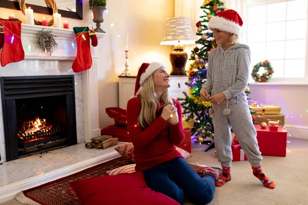 圣诞节时 一个年轻的白种妇女坐在地板上 她的小儿子站在客厅里 戴着圣诞老人的帽子 面带微笑 — 图库照片