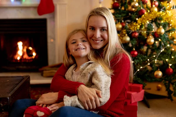 圣诞节时 一位年轻的白种妇女在客厅里用膝盖拥抱着年幼的女儿 在一棵装饰的圣诞树旁 微笑着对着镜头 — 图库照片