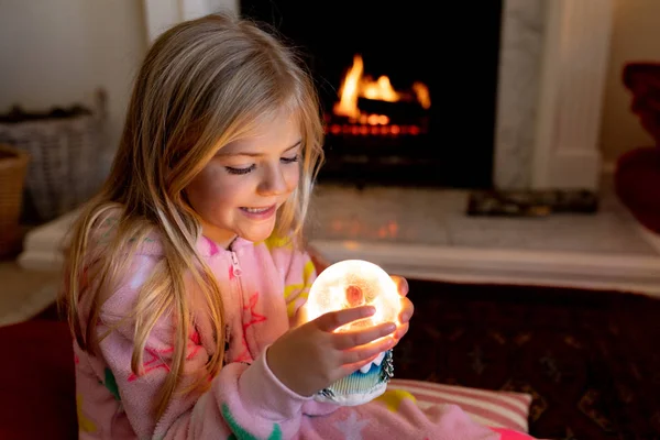 圣诞节时 一个快乐的年轻白种女孩坐在地板上 拿着雪球 在客厅的壁炉旁微笑 — 图库照片