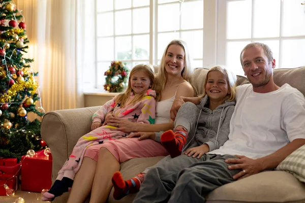 圣诞节时 一对白种人夫妇坐在坐在自己的客厅里 带着年幼的儿子和女儿坐在自己的客厅里 拥抱着 微笑着对着镜头 背景是一棵装饰的树 — 图库照片