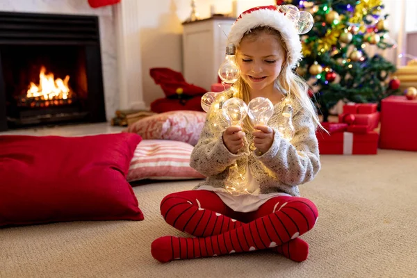 圣诞节时 一个快乐的年轻白种女孩戴着圣诞老人的帽子 在客厅里拿着仙女灯 — 图库照片