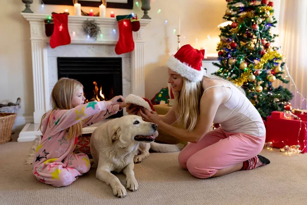 圣诞节时 一个快乐的年轻白种妇女坐在地板上 带着小女儿和他们的宠物狗坐在客厅里 戴着圣诞老人的帽子 背景是装饰的Chritsmas树 — 图库照片