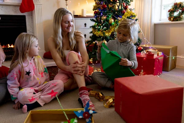 若い白人の母親が クリスマスの時に居間で幼い息子と娘と一緒に床に座り プレゼントを開いて微笑んでいる — ストック写真