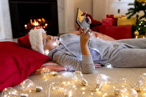 圣诞节时 一个快乐的白人小男孩躺在地板上 戴着圣诞老人的帽子 在客厅里用平板电脑的侧面观看 — 图库照片