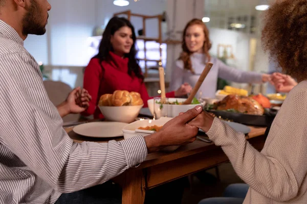 一緒に自宅で感謝祭の夕食を食べる前に 手をつないでテーブルの周りに座っている若い大人の多民族の男女の友人のグループの肩の上に — ストック写真