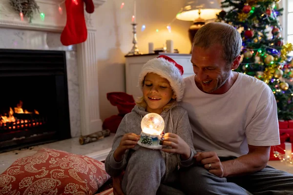 幸せな中年の白人の父親が クリスマスの時に居間で 雪の球体を持ち サンタの帽子をかぶって床に座っている幸せな中年の父親の正面図 — ストック写真