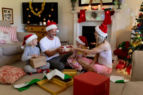 サンタの帽子をかぶり プレゼントを開き 笑顔を見せたクリスマスの時に 幼い息子と娘と一緒に床に座っている白人カップルの側面図 — ストック写真