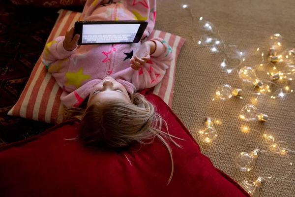 クリスマスの時に彼女の居間でタブレットを使用して床のクッションに横たわっている幸せな若い白人の女の子の正面図 — ストック写真