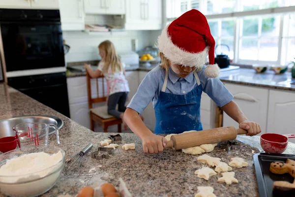 圣诞节前夕 年轻的白人男孩带着妹妹在厨房的工作台上做饼干 一边在厨房里翻滚着面团 — 图库照片