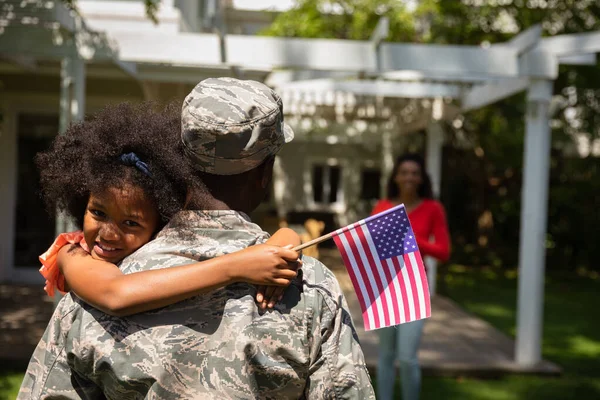 一个年轻的非洲裔美国男性士兵背着他的女儿站在自家花园外 背对着摄像机 举着美国国旗 微笑着 她的母亲在他的身后清晰可见 — 图库照片