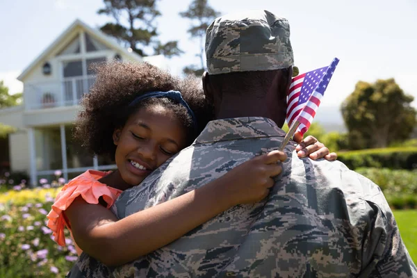 一位年轻的非裔美国士兵背着他的女儿站在自家门外的花园里 紧紧地盯着他 女儿紧靠着他的肩膀 闭着眼睛 举着一面美国国旗 微笑着 — 图库照片