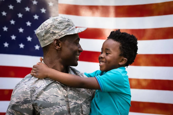 一个年轻的非裔美国士兵背着他年幼的儿子 面面相觑 微笑着 站在一面美国国旗前 侧视近景 — 图库照片