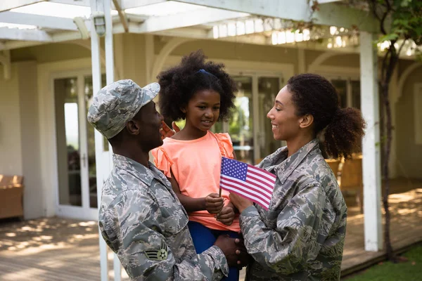 一个年轻的非洲裔美国男子和一个年轻的混血儿站在自家门外的花园里 抱着并拥抱着他们的小女儿 面带微笑 女儿举着美国国旗 — 图库照片