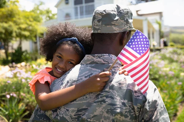 一位年轻的非洲裔美国士兵背着他的女儿站在自家花园的近旁 背对着摄像机 举着美国国旗 面带微笑 — 图库照片