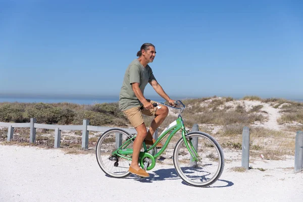 在阳光明媚的日子里 一位年长的白人男子在海滩上享受时光 他骑着一辆背景为沙子的自行车 — 图库照片