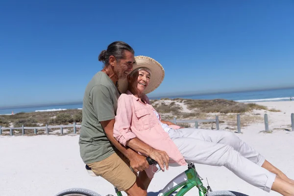 在阳光明媚的日子里 白种人的一对老夫妻在海滩上享受时光 他们骑着一辆自行车 背景是沙子 — 图库照片