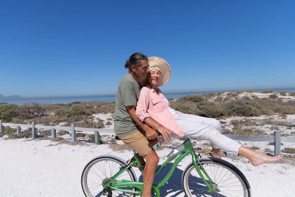 在阳光明媚的日子里 白种人的一对老夫妻在海滩上享受时光 他们骑着一辆自行车 背景是沙子 — 图库照片