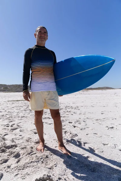 晴れた日にビーチで時間を楽しんでいるシニア白人男性 背景に砂とサーフボードを立って保持します — ストック写真