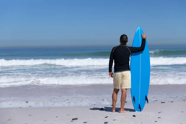 在阳光明媚的日子里 一位年长的白人男子在海滩上享受着时光 他站在海滩上 背对着一只大海的冲浪板 — 图库照片