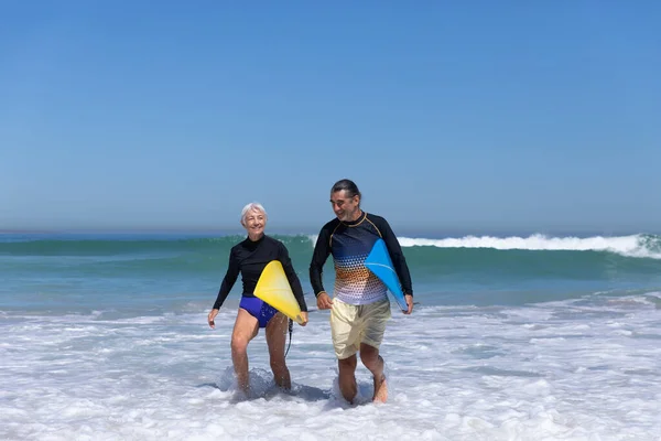 晴れた日にビーチで時間を楽しむシニア白人のカップル 背景に海と歩いてサーフボードを保持します — ストック写真