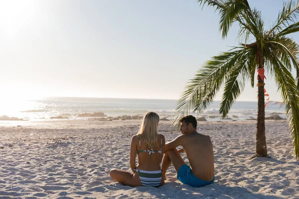 白人夫婦は晴れた日にビーチで時間を楽しみ 背景に海のあるヤシの木に座っています — ストック写真