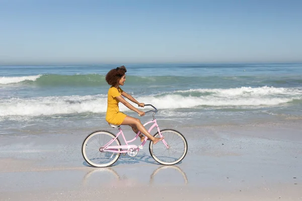 在阳光灿烂的日子里 一位快乐 迷人的混血女子在海滩上享受着闲暇时光 身穿黄色衣服 骑自行车 脸上闪烁着阳光 — 图库照片