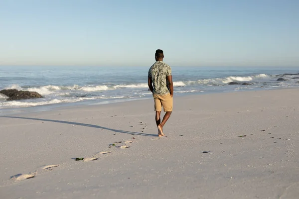 晴れた日にビーチで自由な時間を楽しむ魅力的なアフリカ系アメリカ人の男性の背面ビュー ハワイのシャツを着て 彼に輝くビーチの太陽の下を歩いて — ストック写真