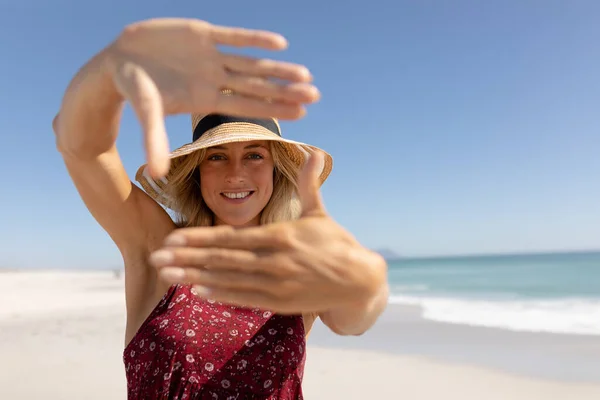 青い空と海を背景に 彼女の顔と笑顔をフレームするために彼女の手を保持し 写真のポーズ 晴れたビーチで自由な時間を楽しんで 休日にわら帽子をかぶってブロンド白人女性 — ストック写真